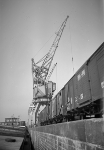 849771 Afbeelding van de overslag van goederen van trein naar schip in de haven te Rotterdam, met behulp van een havenkraan.